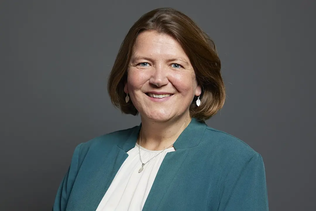 Ellie Chowns, smiling, with shoulder-length brown hair, in a teal jacket over a white top, against a grey background