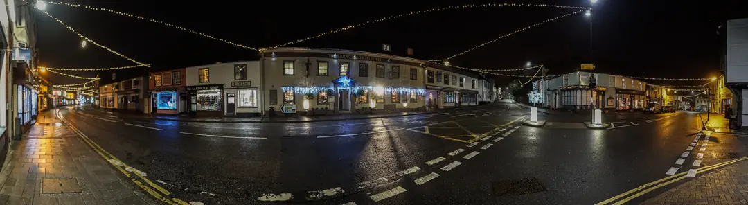 A night panorama of Great Dunmow’s Market Place and High Street under strings of Christmas lights: rows of lit shopfronts and the Saracen’s Head hotel, decorated with blue lights and a star, around a wet, empty junction
