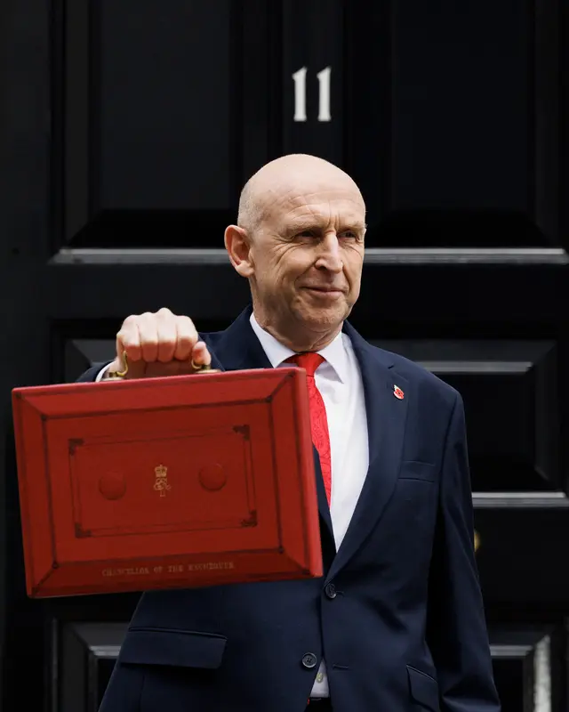 The Chancellor, John Healey, smiling outside the black door of 11 Downing Street, holding up the red Budget box marked “Chancellor of the Exchequer”